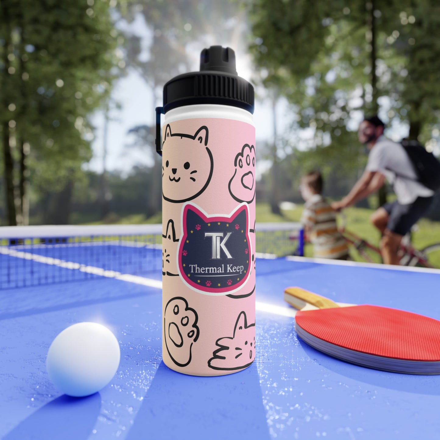 Pink water bottle with cat design on a table tennis table, with people playing in the background.